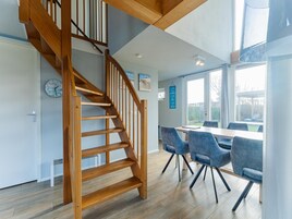 Un interior moderno con una escalera de madera, un comedor con sillas azules y vistas amplias a través de grandes ventanales.