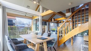 Ein modernes Interieur mit einer Holztreppe, einem Essbereich mit Tisch und Stühlen sowie großen Fenstern mit Außenblick.