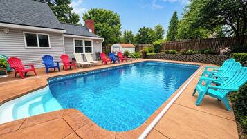 Une piscine avec une terrasse carrelée, entourée de fauteuils Adirondack, et une maison en arrière-plan.