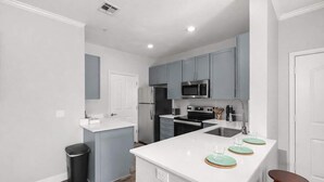 A modern kitchen with grey cabinets, white countertops, and stainless steel appliances. There is a trash can and a small island with stools. - Scottsdale Desert Escape 2BR with Sunrise Peak Views, Pets OK (Scottsdale)