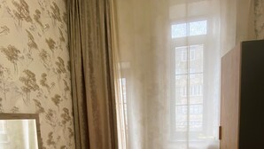 A bedroom with a wooden dresser, a bed with a polka dot bedspread, a window with curtains, and floral wallpaper.