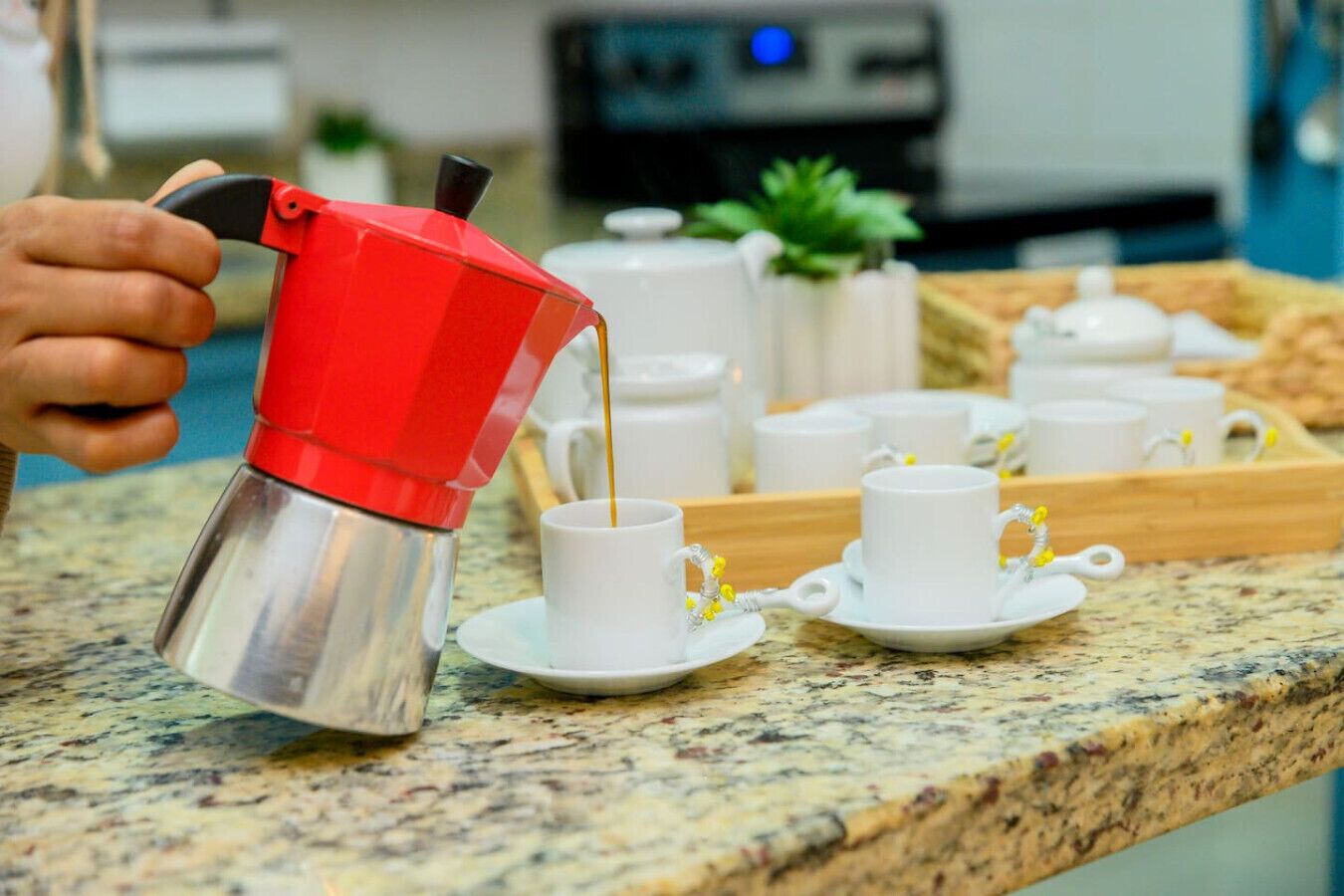 Une personne verse du café d’une cafetière rouge et argentée dans une tasse blanche posée sur un comptoir en granit.