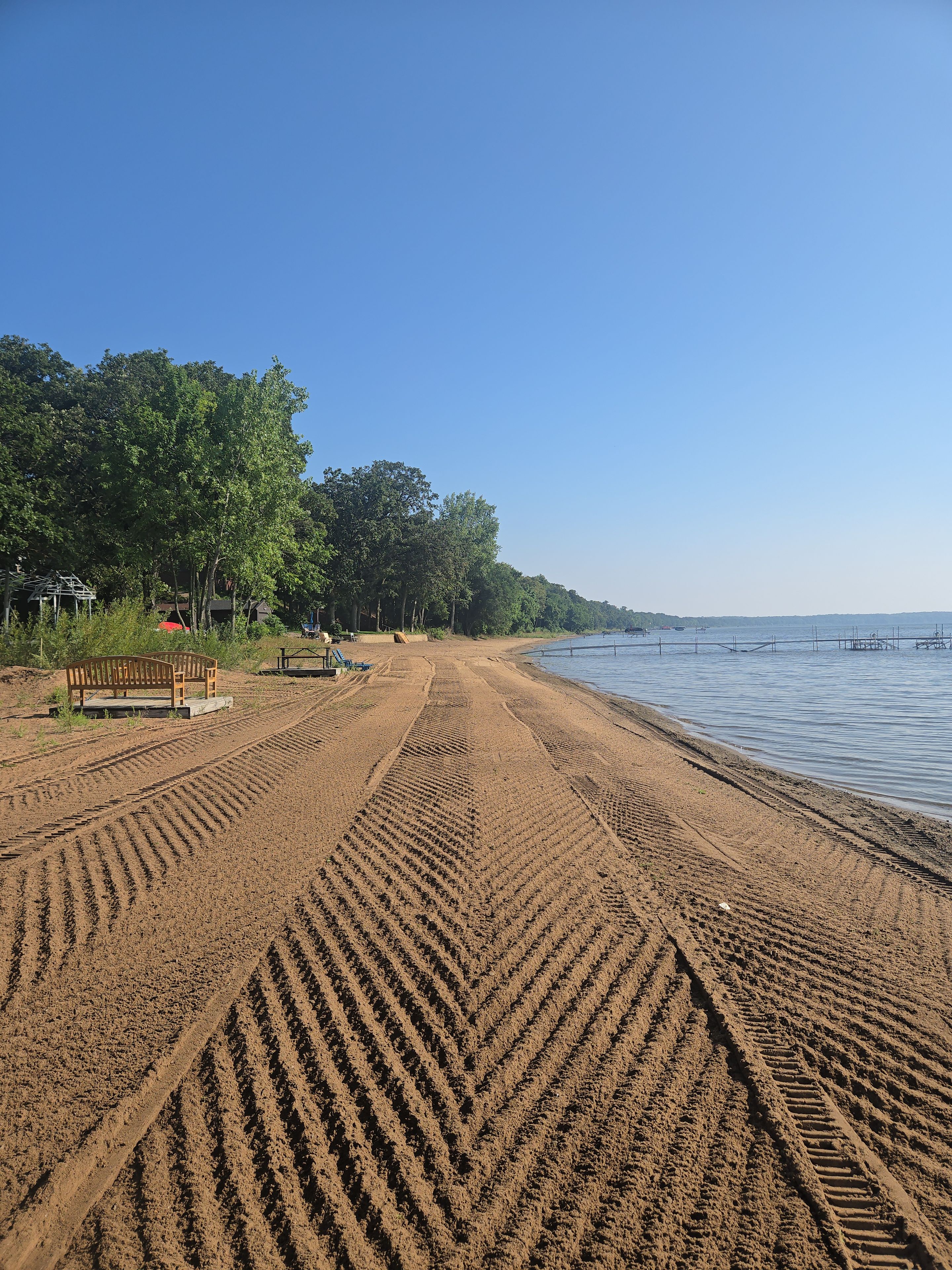 A sandy beach with a wooden bench and a small structure, likely a gazebo, surrounded by trees.