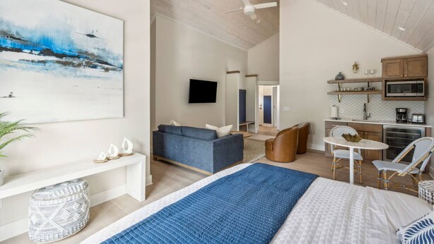 Una camera d'albergo moderna con un letto grande, un tappeto blu, un divano, un tavolo da pranzo con sedie, un angolo cottura e una TV a parete.