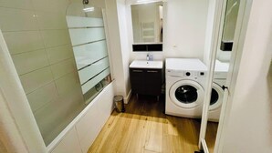 Casa de banho compacta com cabine de duche em vidro, lavatório, máquina de lavar e espelho.