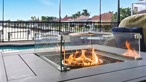 A glass-enclosed fire pit with flames, a pool, and a view of palm trees and buildings in the background. - Sunset Retreat Pool/Spa, great views, we love dogs (Punta Gorda)