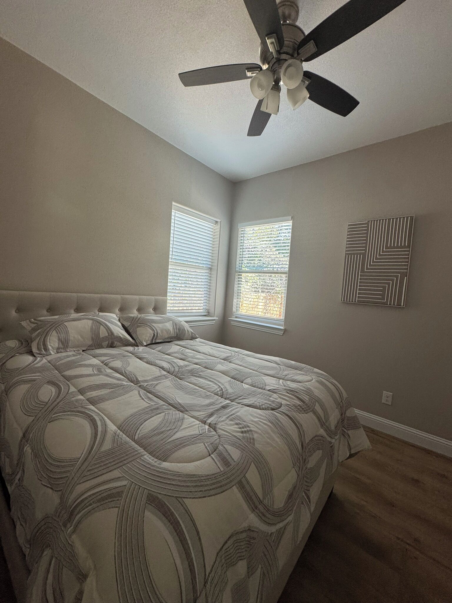 A bedroom with a tufted headboard, a bed with a patterned bedspread, a ceiling fan, two windows with blinds, and a geometric wall art piece.