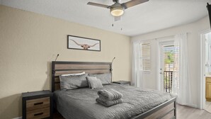 Ein Schlafzimmer mit Bett, Nachttischen, Deckenventilator, einem gerahmten Bild und Blick durch ein Fenster mit Jalousien.