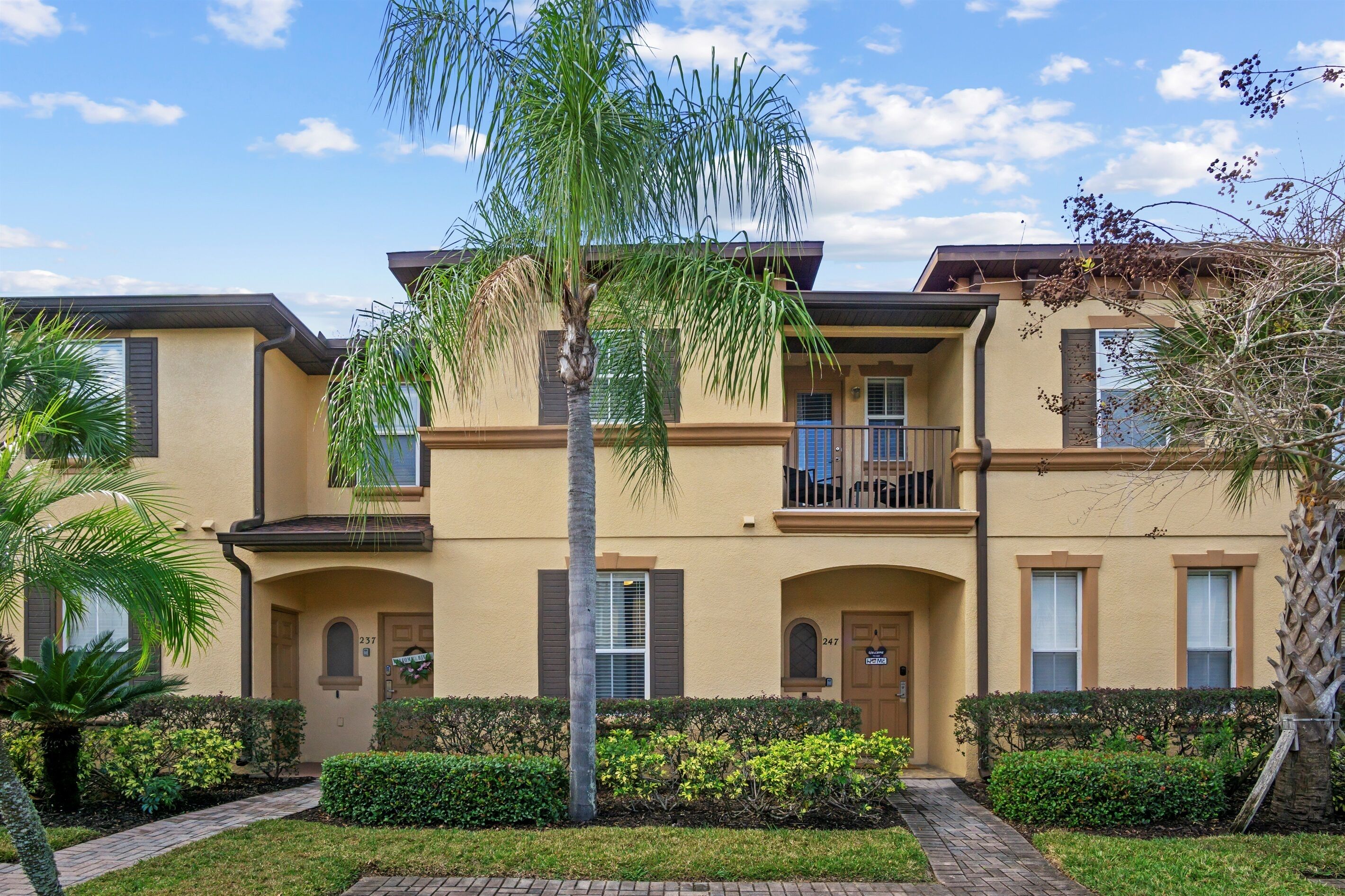 A multi-story residential building with a beige exterior, balconies, and a well-maintained garden with palm trees and shrubs.