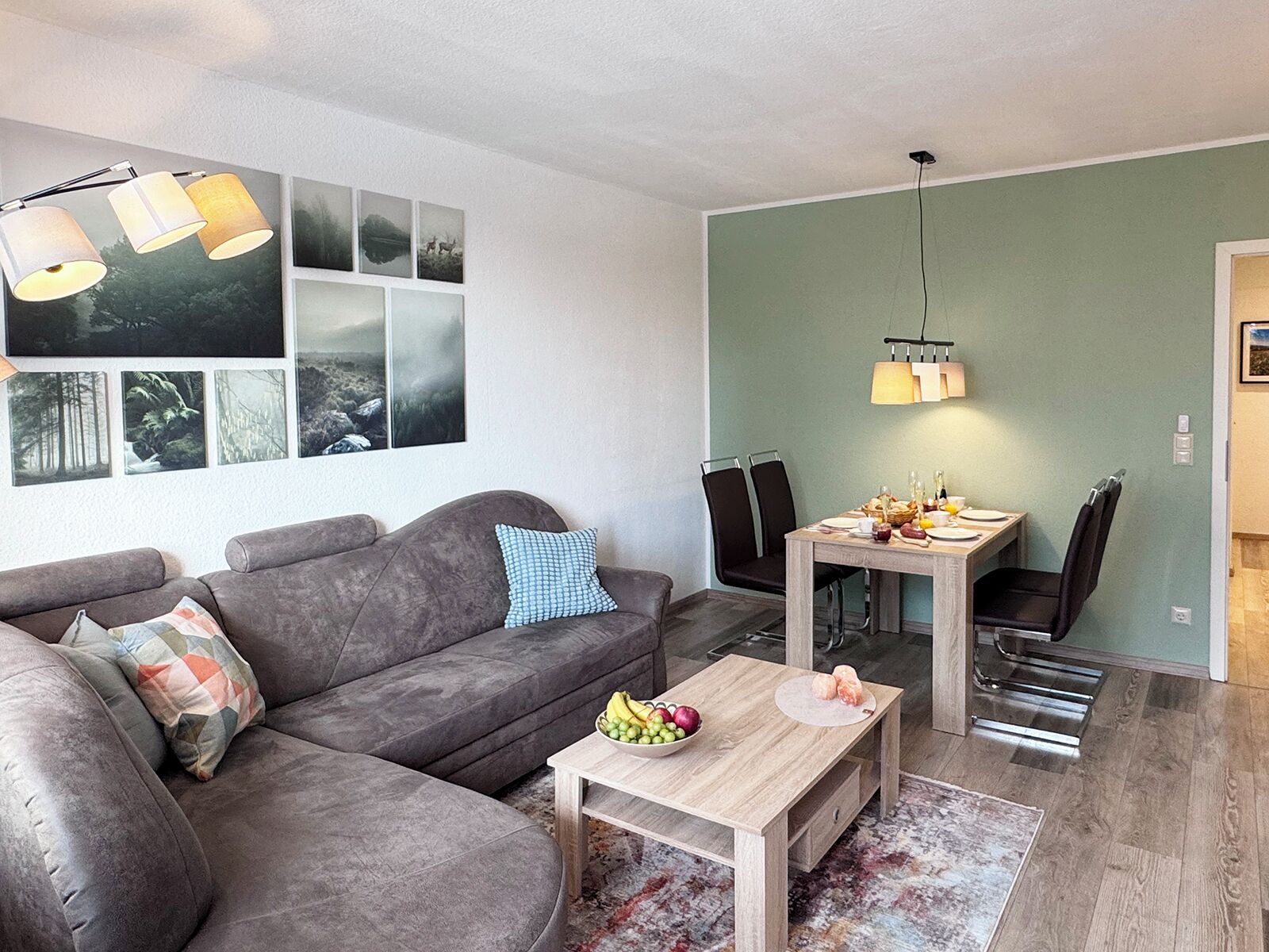 A modern living room with a grey sofa, a wooden coffee table with fruits, and a dining area with a table and chairs.