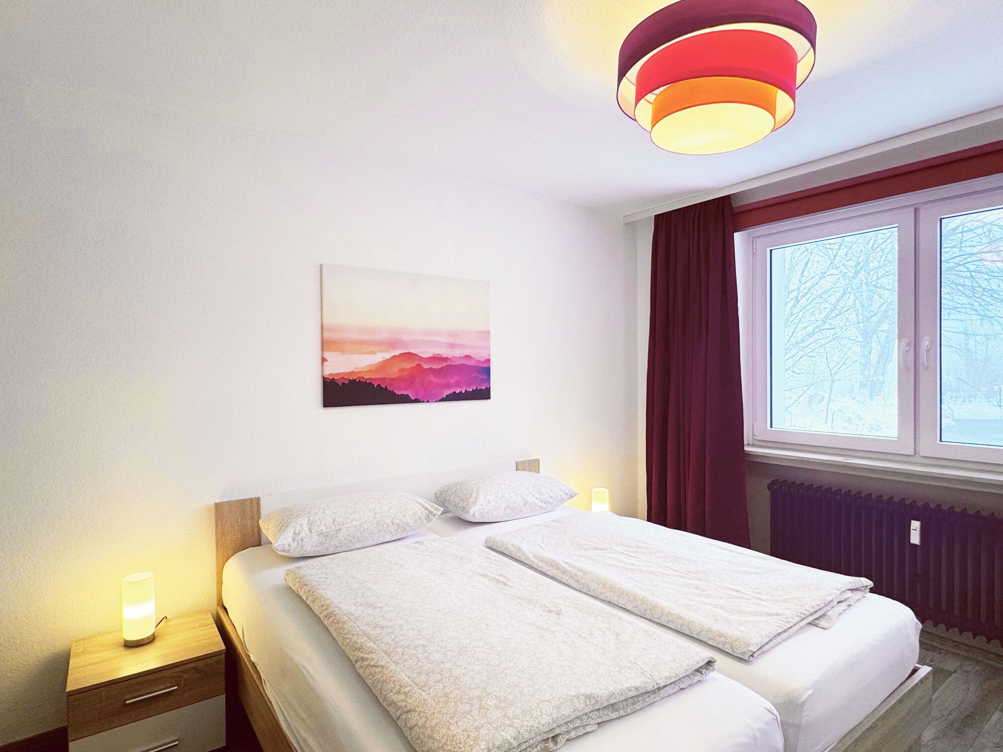 A hotel room with a bed, a nightstand, a painting, a window with curtains, and a ceiling light fixture.