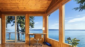 Eine Holzhütte mit Schreibtisch und Stuhl, große Fenster mit Blick auf Wasser und Bäume.