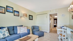 A living room with a blue sofa, a matching armchair, a coffee table, and a dining area with a white table and chairs. - Coastal Chic 1BR Oceanfront Suite (Myrtle Beach)