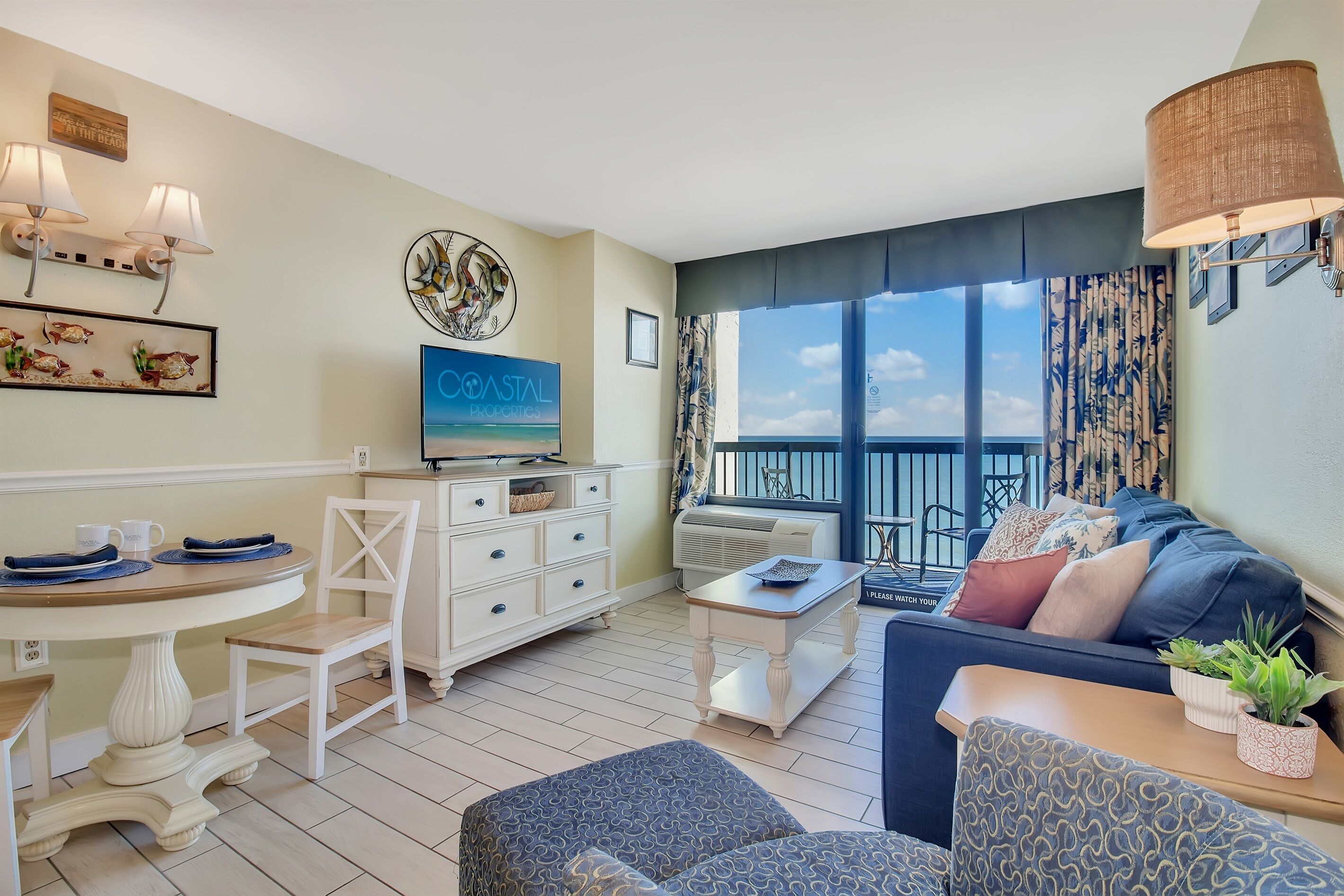 A modern living room with a TV, white furniture, a balcony with a view, and coastal decor.