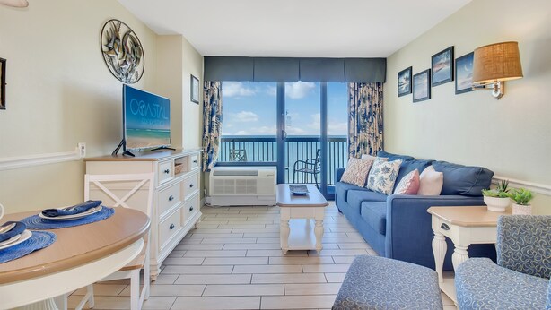 A modern living room with a sofa, a dining table, and a TV. There is a balcony with a view of the sky and a railing. - Coastal Chic 1BR Oceanfront Suite (Myrtle Beach)