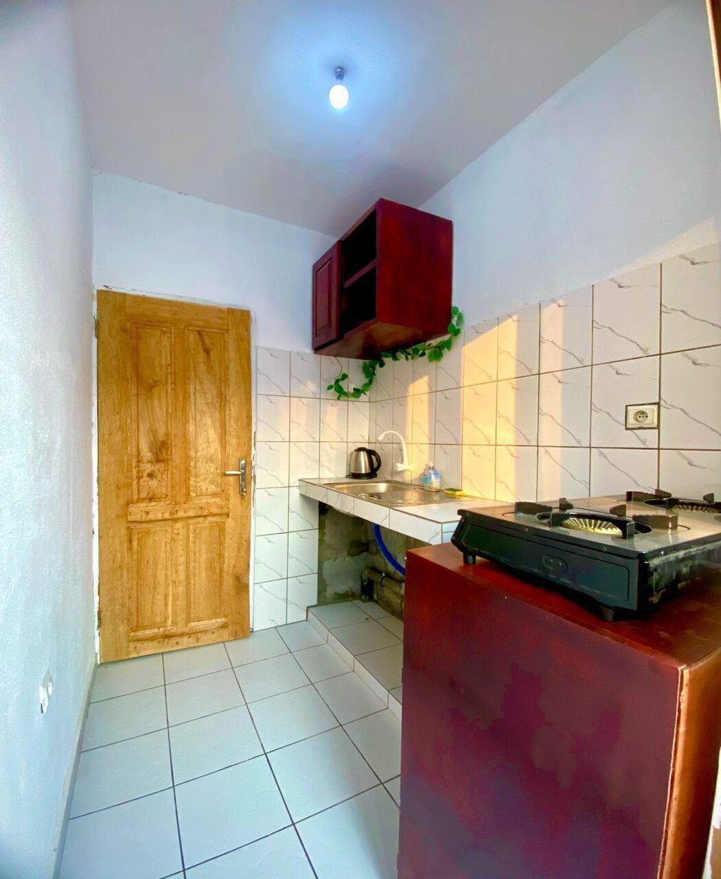 A compact kitchen with a wooden door, white tiled walls, a red cabinet, and a gas stove.
