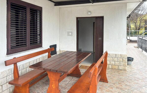 Een terras met een houten tafel en banken, een raam met zonwering en een deur naar binnen.