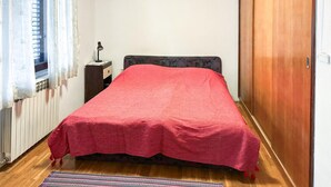 Ein Schlafzimmer mit einem Bett, das mit einer roten Decke bezogen ist, einem Holzboden, einem Heizkörper, einem Fenster mit Vorhängen, einem Nachttisch mit Lampe und einem Holzschrank.