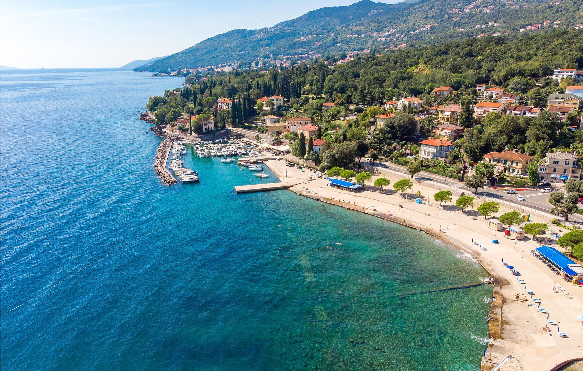 An aerial view of a coastal town with a marina, clear blue waters, and a mix of residential buildings.
