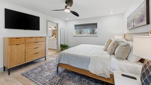 A bedroom with a bed, a wooden dresser, a ceiling fan, a TV, and a window with a view of trees.