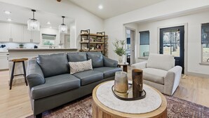 A spacious living area with a grey leather sofa, a round coffee table, and a white armchair. A wooden bookshelf and a small side table are visible in the background.