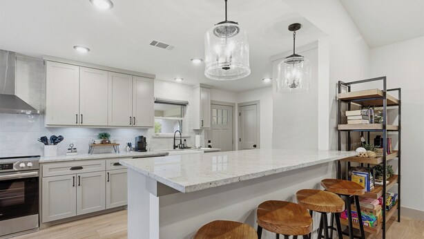 A modern kitchen with white cabinetry, a large island with stools, pendant lighting, and a metal bookshelf with books.
