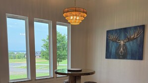 Ein Zimmer mit einem runden Tisch, Hockern mit Geweihbeinen, einer Chandelierleuchtre, einem Kuhfellteppich und einem Elchgemälde.