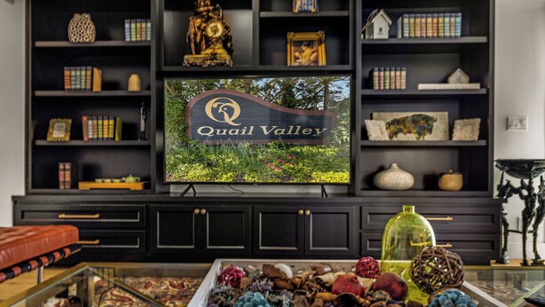 A living room with a television displaying 'Quail Valley' sign, a glass coffee table with decorative items, and a bookshelf with various objects. - Farm barn with territorial view in Banks w/ pheasants- 1 hour drive to coastal (Banks)
