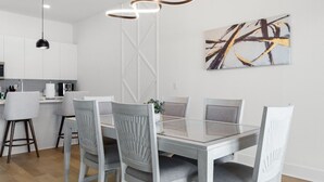 Un área de comedor moderna con una mesa de vidrio y sillas grises tapizadas, una cocina al fondo y un cuadro abstracto contemporáneo en la pared.