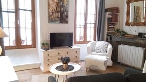 A living room with a TV, a white sofa, a white armchair, a small round table with a plant, and a bookshelf with books and a mirror. - Charming 26m2 studio in the center, 50m from the thermal baths (Bagnères-de-Bigorre)