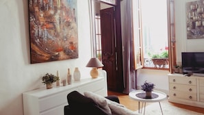 A living room with a large abstract painting, a sofa, a TV, and a window with curtains. - Charming 26m2 studio in the center, 50m from the thermal baths (Bagnères-de-Bigorre)