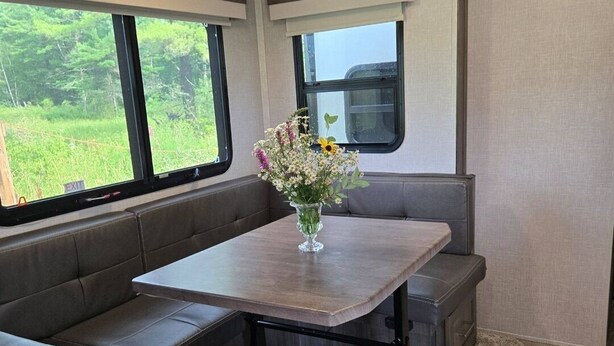 Interni di un camper compatti con zona pranzo, tavolo e sedute a panca, un piccolo oblò che affaccia sul verde e una lampada a soffitto.