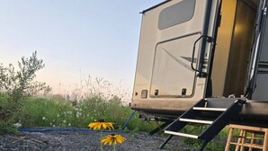 Un camper con porta scorrevole, un piccolo tavolo di legno e un'area giardino con rocce e fiori gialli.