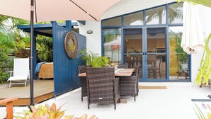 En udendørs terrasse med fletningsmøbler, en blå væg og en glasdør, der fører til et køkken.