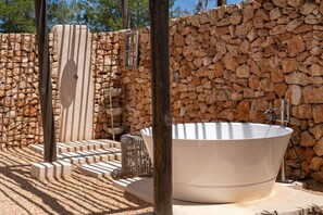 A stone-walled outdoor spa area with a large freestanding bathtub, a wooden bench, and a small table with a bowl. -  New Finca Samavara 6 bedrooms Holistic luxury retreat in Ibiza (SANTA EULARIA DES RIU)