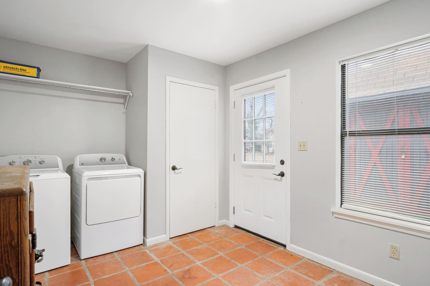 A laundry room with two white washing machines, a white door, and a window with blinds.