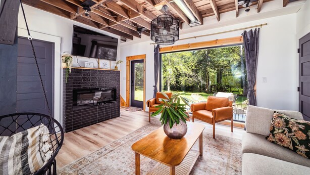 A modern living room with a fireplace, wooden ceiling beams, a large window, and a view of the outdoors. - Renovated Barn w/ Jacuzzi - Tree Table - 3 Acres (Ruckersville)