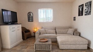Un salon avec un canapé, un téléviseur posé sur un meuble, un pouf en osier et une fenêtre avec des rideaux.