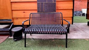 Un banco de metal negro con asiento y respaldo de listones, ubicado sobre césped artificial frente a una pared de madera con un detalle de piedra.