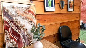 Una pared de madera con un cuadro abstracto enmarcado, un cartel enmarcado de mojito, un cartel enmarcado de margarita, un jarrón blanco con hojas verdes y una silla negra.