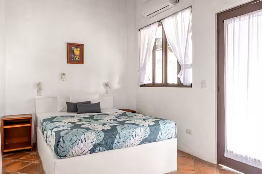Une chambre à coucher comprenant un lit, une table de chevet, une fenêtre avec des rideaux et un climatiseur fixé au mur.