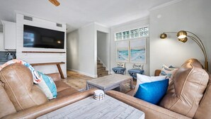 A living room with a leather sofa, a coffee table, a TV mounted on a white cabinet, and a staircase leading to another room. - Steps to Beach & Ocean View | Good Morning Rentals (Jacksonville Beach)