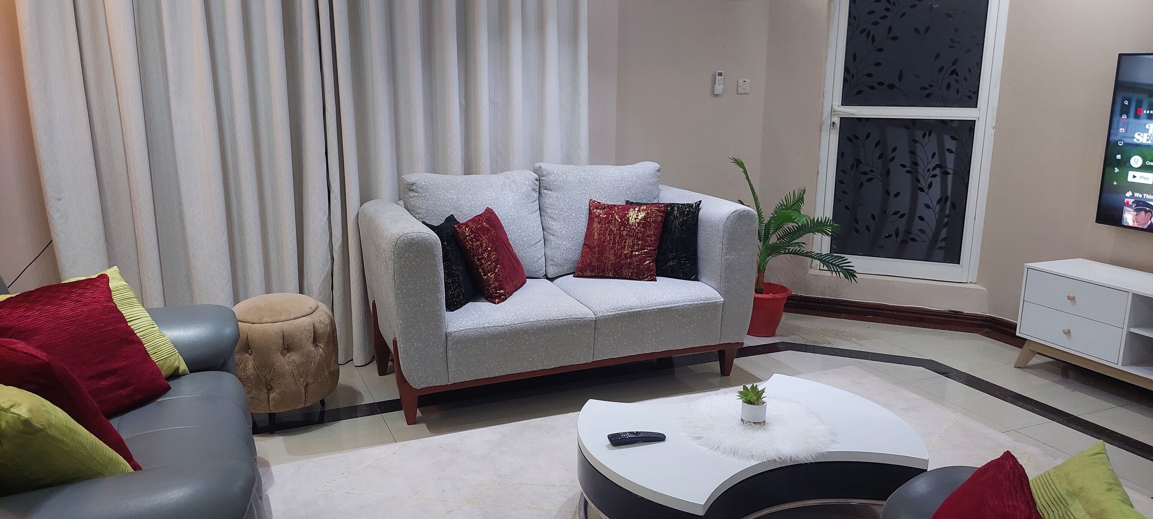 Un salon avec un canapé gris, un petit pouf rond, une table basse blanche ornée d’une plante et un téléviseur fixé au mur.