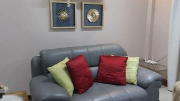 Un canapé en cuir gris agrémenté de coussins rouges et verts, un petit tapis blanc et moelleux, et deux éléments de décoration encadrés accrochés au mur.