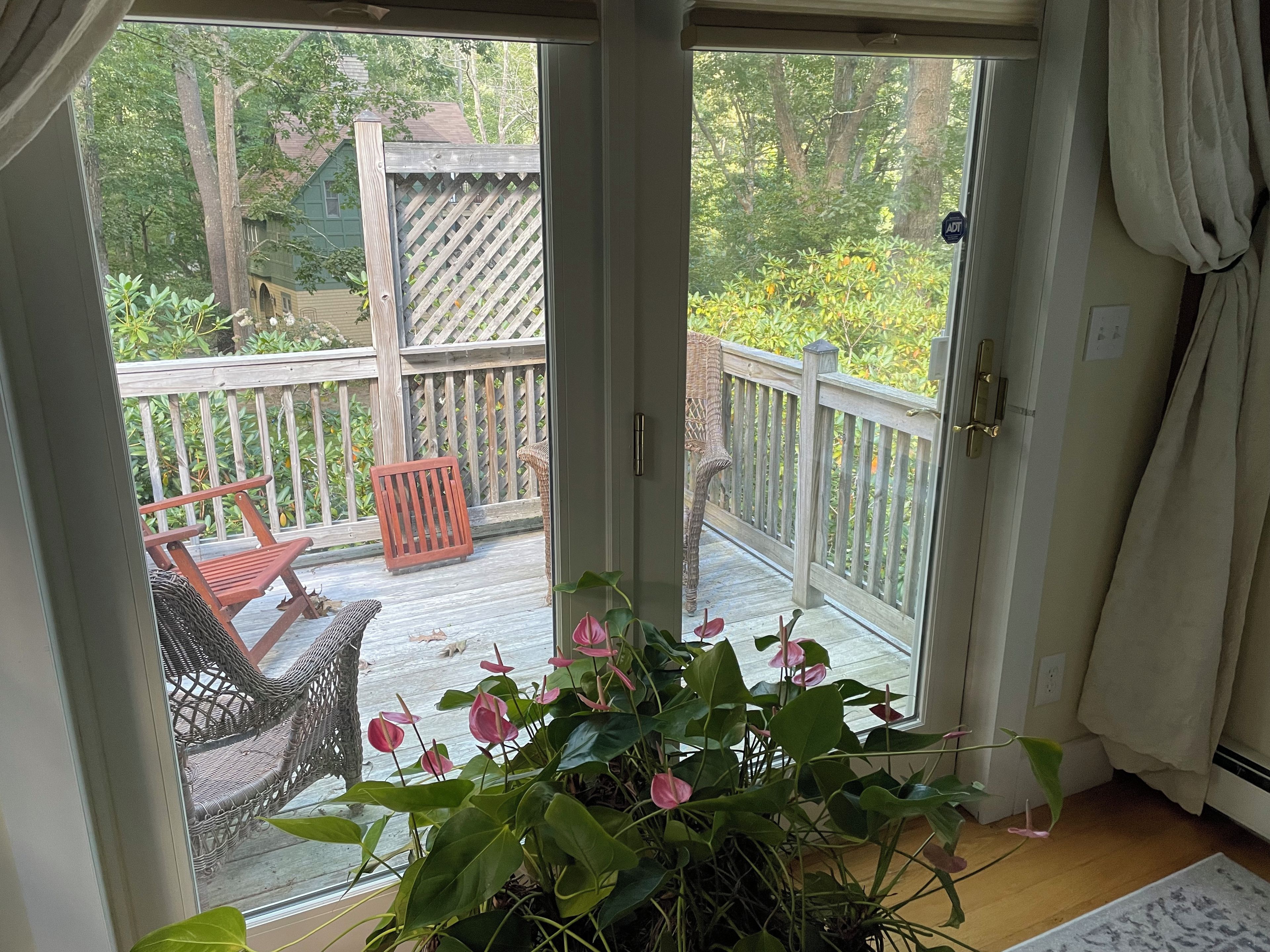 Une porte-fenêtre coulissante donne sur une terrasse extérieure agrémentée d'une rambarde en bois, de chaises en osier et d'un pot de fleurs aux fleurs roses.