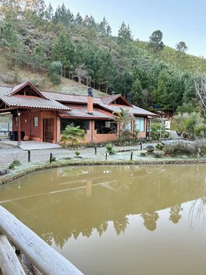 A rustic cabin with a tiled roof, surrounded by a small pond and dense forest.