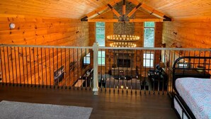 A spacious cabin with a high ceiling, wooden walls, a central fireplace, and a balcony with a railing.