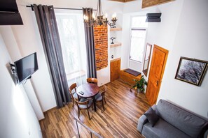 A cozy living room with a grey sofa, a wooden dining table with chairs, a TV mounted on the wall, and a brick wall with a chandelier. - Lovenovsky Apartments (Krakow)