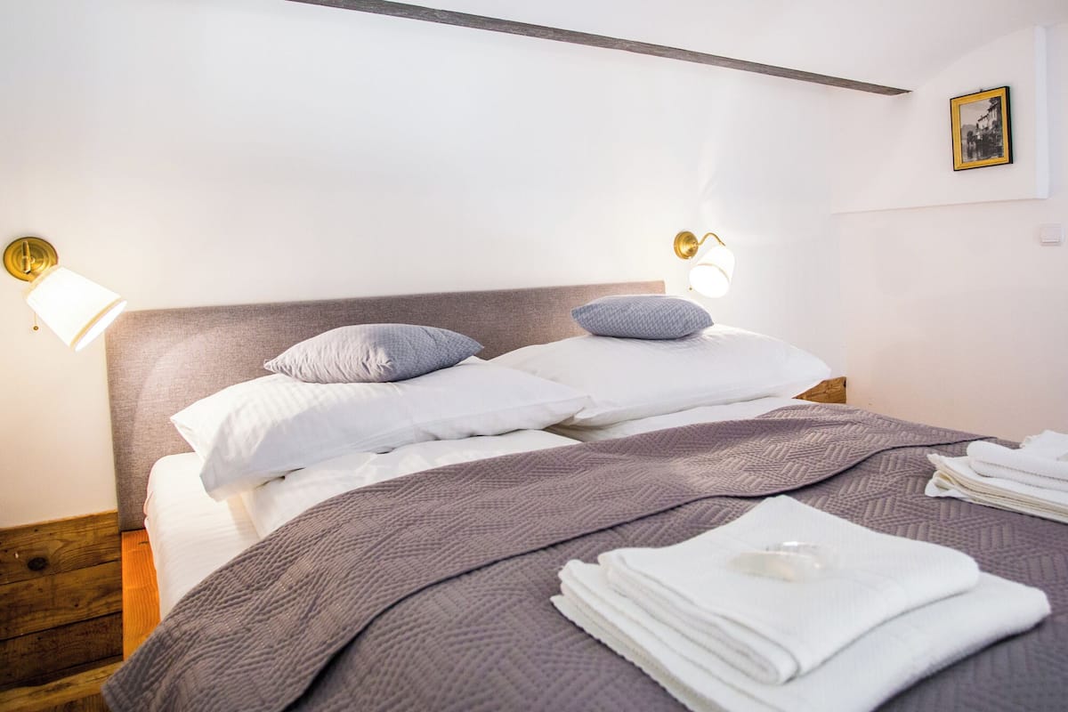 A neatly made bed with a grey quilt, white pillows, and a folded white towel on the side.