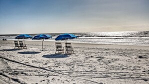 Ein Strand mit drei blauen Sonnenschirmen und zwei leeren Liegestühlen.